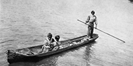 Man poles a small boat through the water while two people sit in it