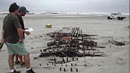 Two archaeologists examining a partially revealed shipwreck on a beach